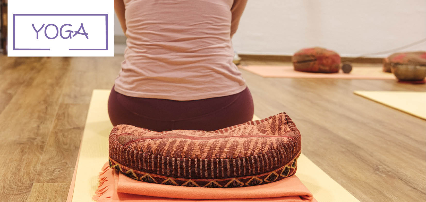 Der untere Rücken einer Frau von hinten, die auf Yogakissen sitzt, links oben der Text: YOGA